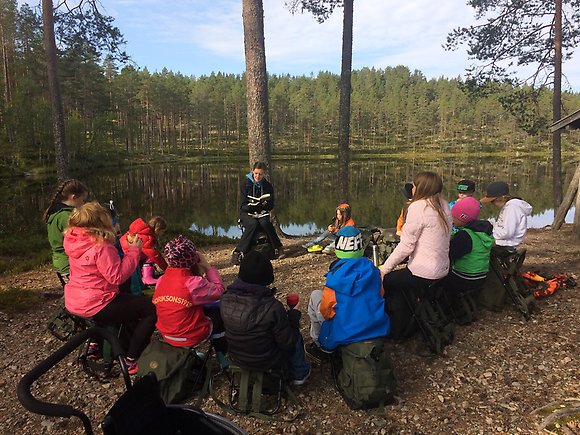 Elever som sitter i en ring utomhus vid en sjö med en lärare som läser för dem ur en bok.